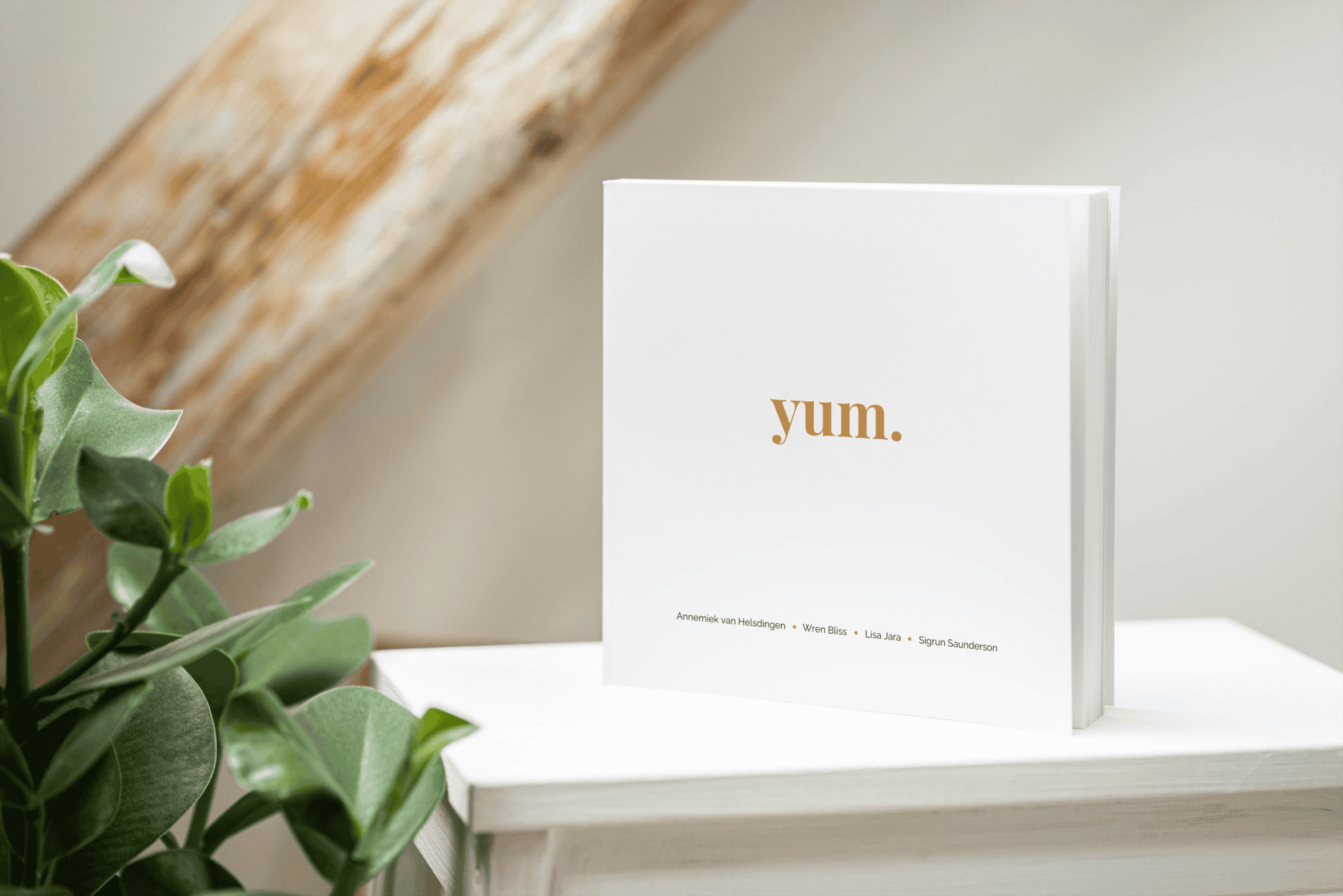 A book with a white cover and the word Yum. in a light golden tan colour stands on a white table, with a plant to its left and a wooden beam in the background, giving the impression that it's in a cosy attic.